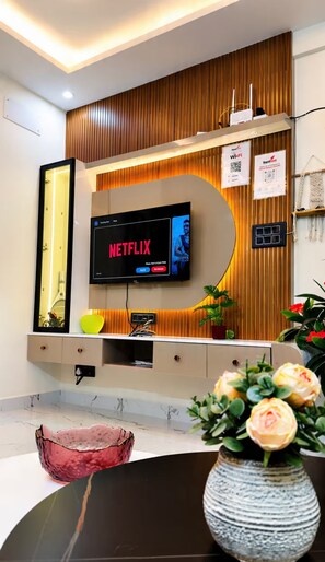 A modern living room with a flat-screen TV mounted on a wooden wall, a round coffee table, and a vase with flowers. - RapidNest - HSR Where Cozy Meets Luxury (Bengaluru)