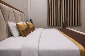 A hotel room with a bed, pillows, a headboard, a window with curtains, and a chair. - Shri Hari Guesthouse (Varanasi)