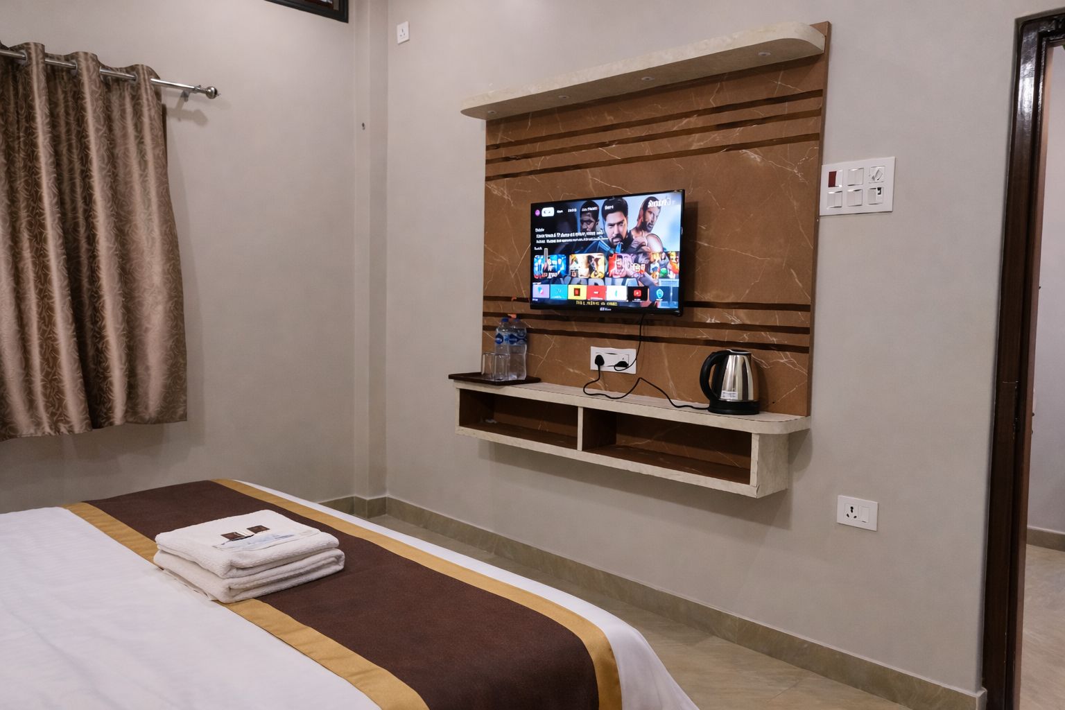 Habitación de hotel con una cama, un televisor montado en un panel de madera, un estante con una tetera y vasos, y una cortina.