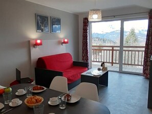 A modern room with a red sofa, dining table, and chairs. There are two wall-mounted lights, a window with a view of mountains, and a small table with a figurine. - Ski aux pieds | Piscine Chauffée | Spa (Combloux)