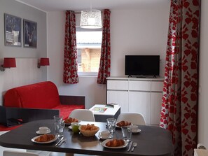 A room with a dining table set for breakfast, a red sofa, a TV, and a window with curtains. - Ski aux pieds | Piscine Chauffée | Spa (Combloux)