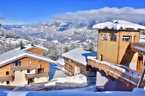 A wooden chalet with a balcony, surrounded by snow-covered mountains and a clear sky. - T2 au Pied des Pistes | Espace bien-être (Combloux)