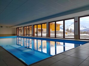 An indoor swimming pool with clear windows, a tiled floor, and a view of a mountain landscape. - Directement sur les pistes | Piscine | Balcon (Combloux)