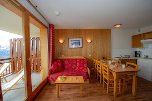 Una habitació acollidora amb un sofà de fusta, una taula de menjador amb cadires, una zona de cuina i un finestral gran amb vistes.