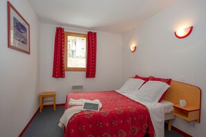 A hotel room with a bed, a bedside table, a window with curtains, a wall-mounted light, and a small stool. - Ski aux Pieds | Piscine Chauffée | Balcon (Saint-François-Longchamp)