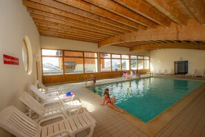 An indoor swimming pool with a wooden ceiling, a view of mountains through large windows, and several white plastic chairs. - Au pied des pistes | Balcon | Piscine Chauffée | (Saint-François-Longchamp)