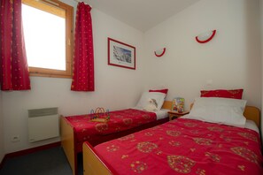 A bedroom with two single beds, a window with red curtains, a framed picture on the wall, and a small bedside table with a book. - Au pied des pistes | Balcon | Piscine Chauffée | (Saint-François-Longchamp)