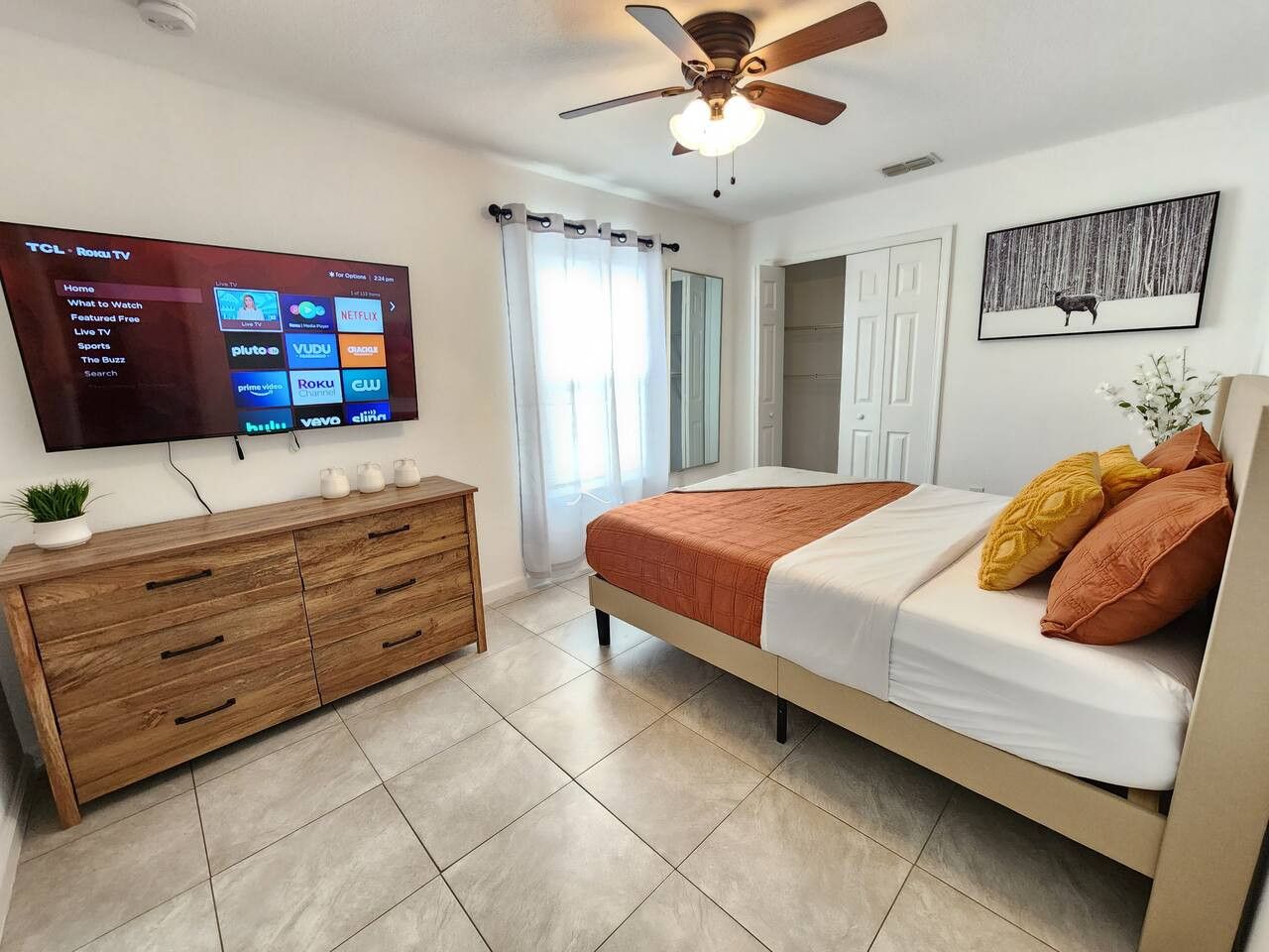 Ein Schlafzimmer mit einem Bett, einem an der Wand befestigten Fernseher, einer Holzkommode und einem Deckenventilator.