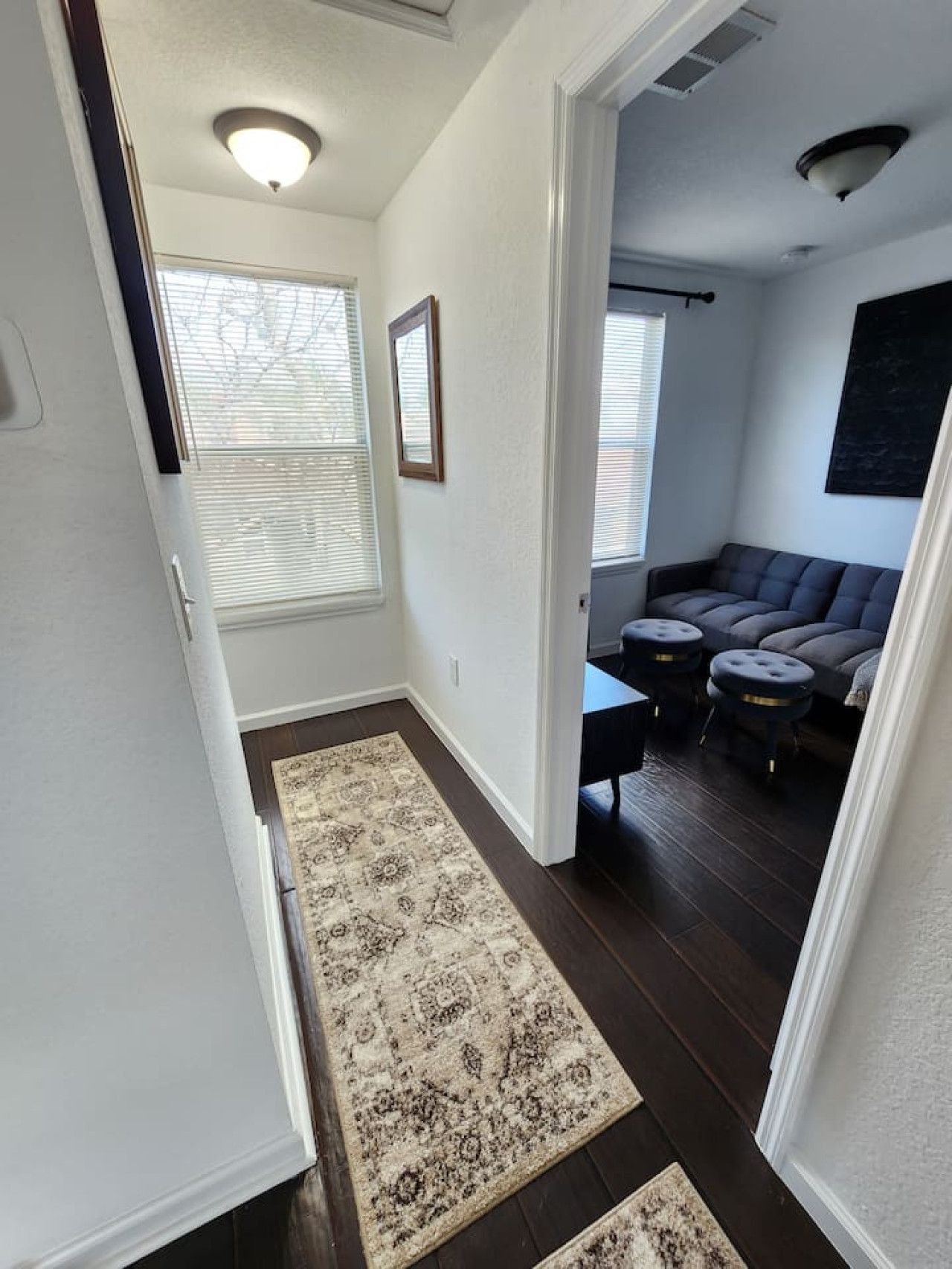 A hallway leading to a living area with a dark wood floor, a patterned rug, and a dark-colored sofa with matching stools.