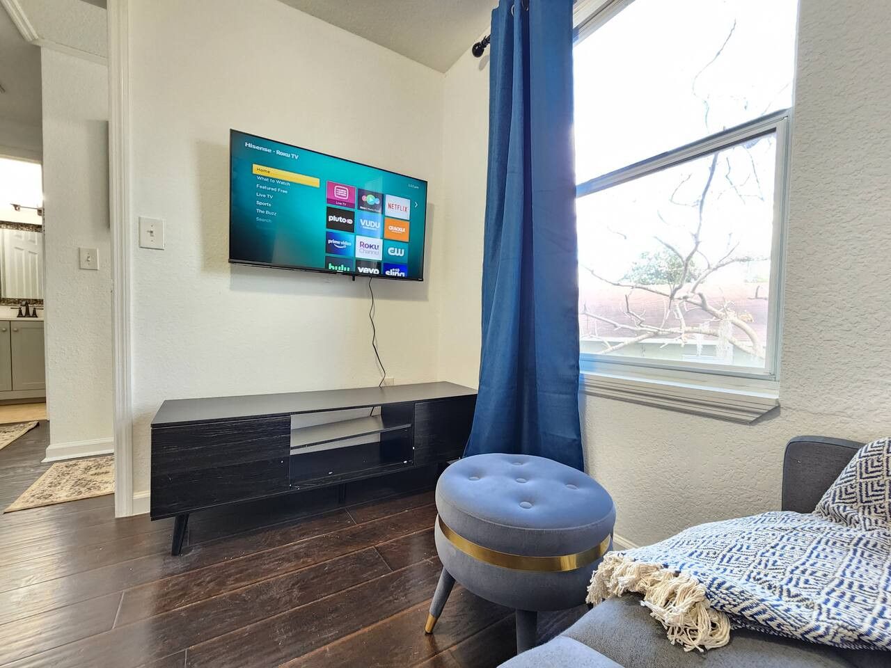 A modern living room with a flat-screen TV mounted on the wall, a dark wooden entertainment unit, a blue curtain, and a patterned throw on a couch.