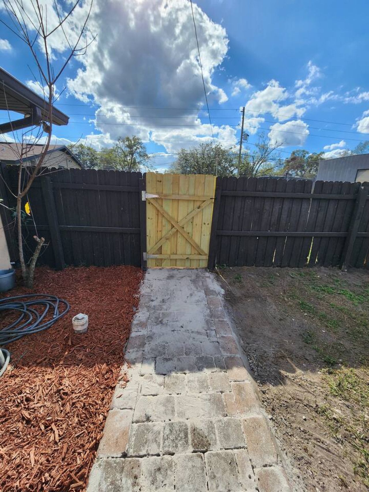 A backyard with a paved pathway leading to a wooden gate, surrounded by a wooden fence, mulch, and a grassy area.