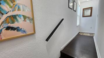 En hall med en vit, strukturerad vägg, en inramad abstrakt målning, ett svart handräcke och ett mörkt trägolv.