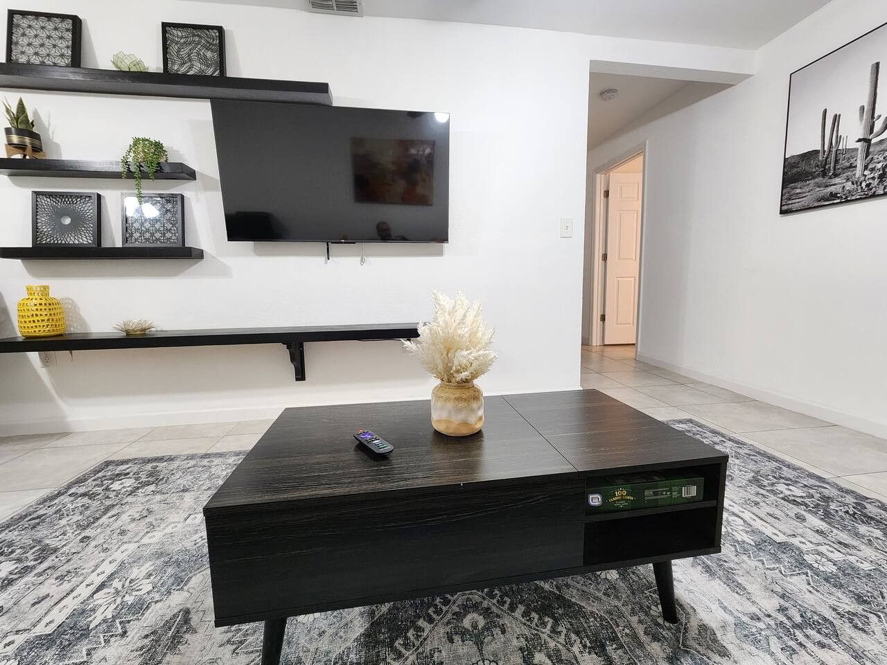 Un salon avec un téléviseur à écran plat fixé au mur, une table basse en bois foncé sur laquelle reposent une télécommande et un vase décoratif, et un tapis à motifs sur le sol.
