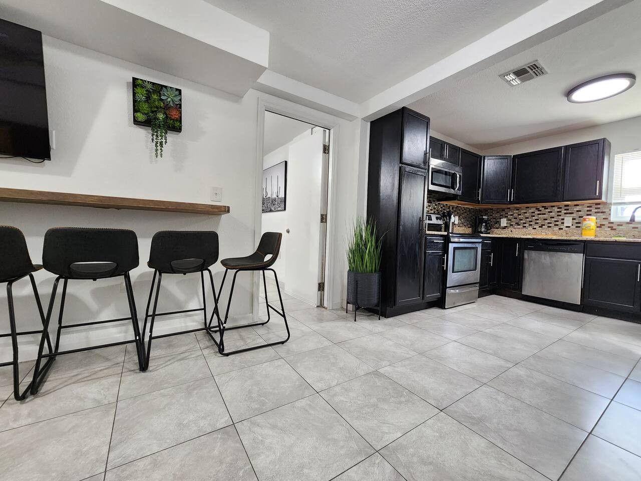 A modern kitchen with dark cabinetry, a stainless steel oven, and a microwave. There are three black bar stools and a potted plant.
