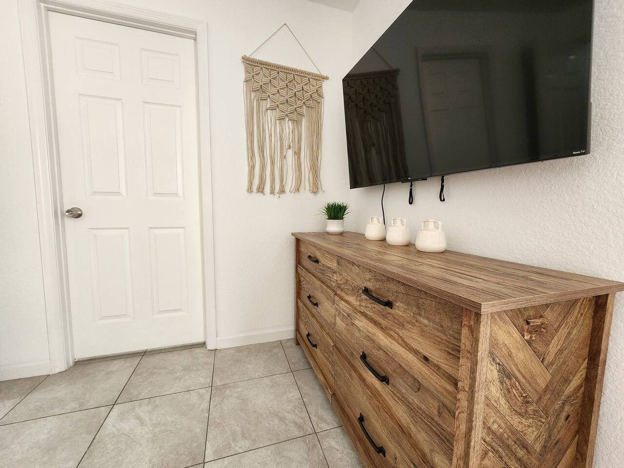 A room with a wooden herringbone-patterned dresser, a flat-screen TV mounted on the wall, and a macramé wall hanging.