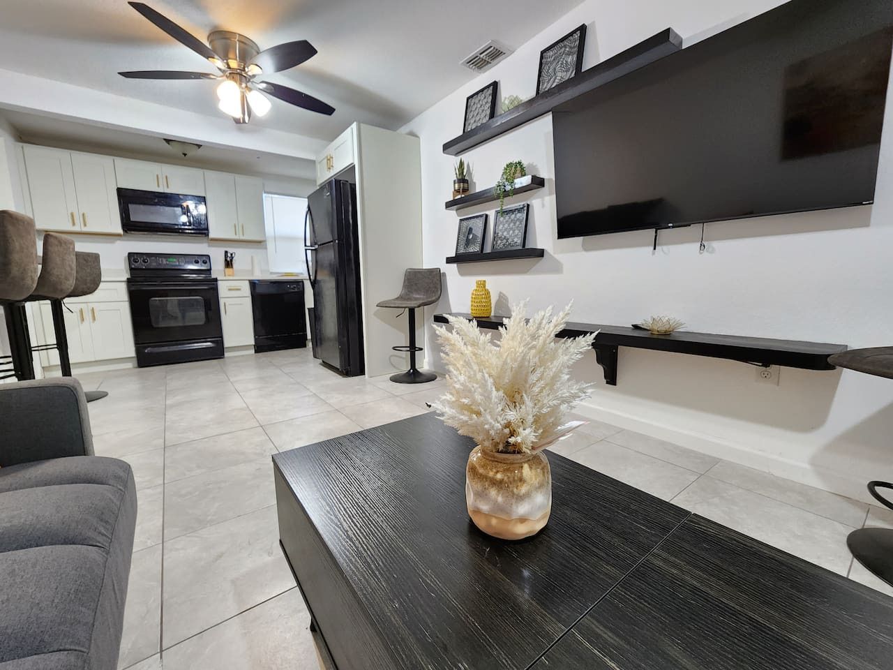 Un salon moderne avec une table basse en bois foncé, un canapé gris et un vase décoratif. On aperçoit, au fond, une cuisine avec des meubles blancs et un réfrigérateur noir.