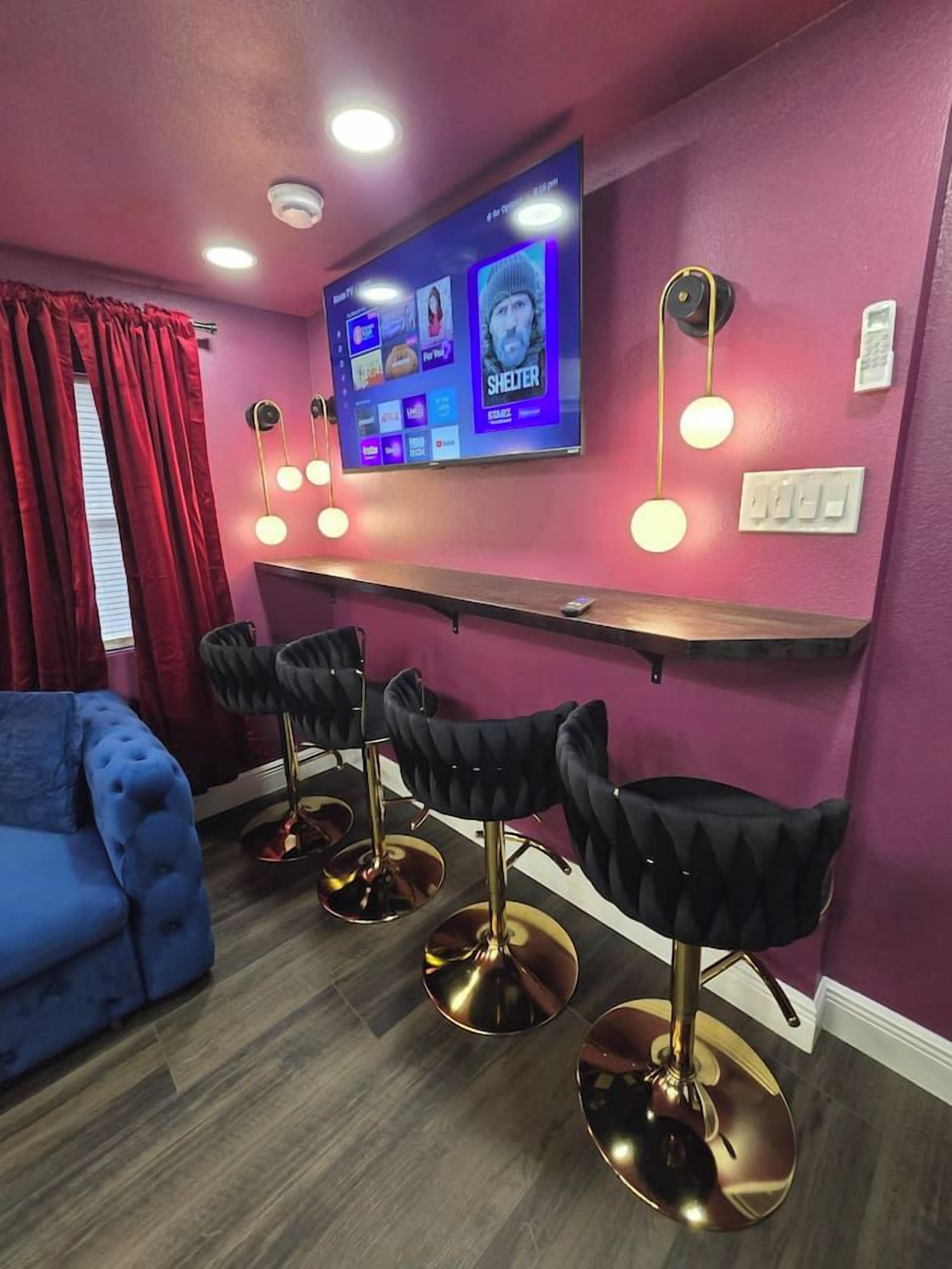 Une pièce avec un canapé bleu, un téléviseur fixé au mur, un coin bar avec des tabourets et un rideau rouge.