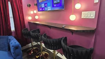 Une pièce avec un canapé bleu, un téléviseur fixé au mur, un coin bar avec des tabourets et un rideau rouge.
