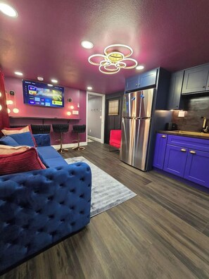 Un salon moderne avec un canapé bleu, un téléviseur à écran plat et un coin cuisine doté de meubles violets.