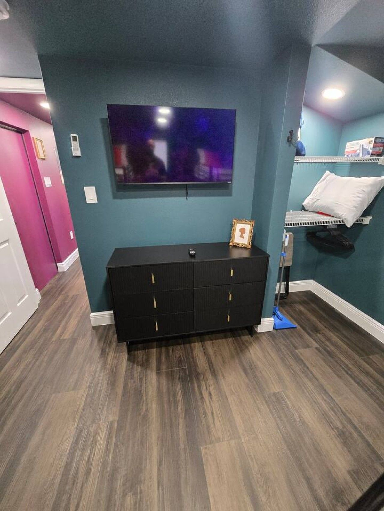 Une pièce dotée d’un parquet en bois foncé, d’un meuble de télévision noir, d’un téléviseur à écran plat fixé au mur et d’un lit avec un oreiller blanc et un tableau encadré.