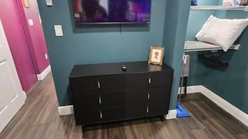 Une pièce dotée d’un parquet en bois foncé, d’un meuble de télévision noir, d’un téléviseur à écran plat fixé au mur et d’un lit avec un oreiller blanc et un tableau encadré.