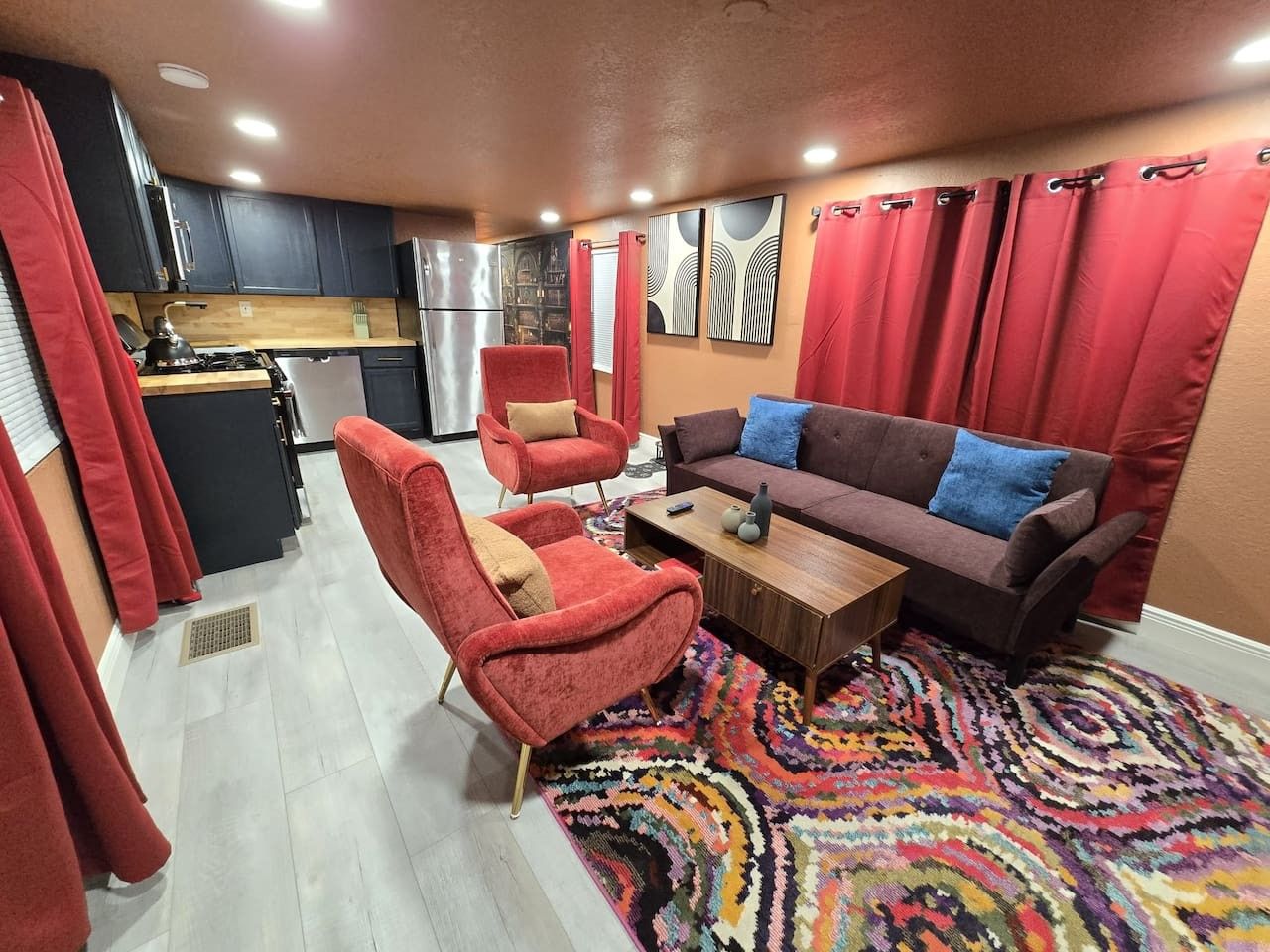 Un espace de vie compact comprenant un coin cuisine, un canapé, un fauteuil rouge et un tapis à motifs.