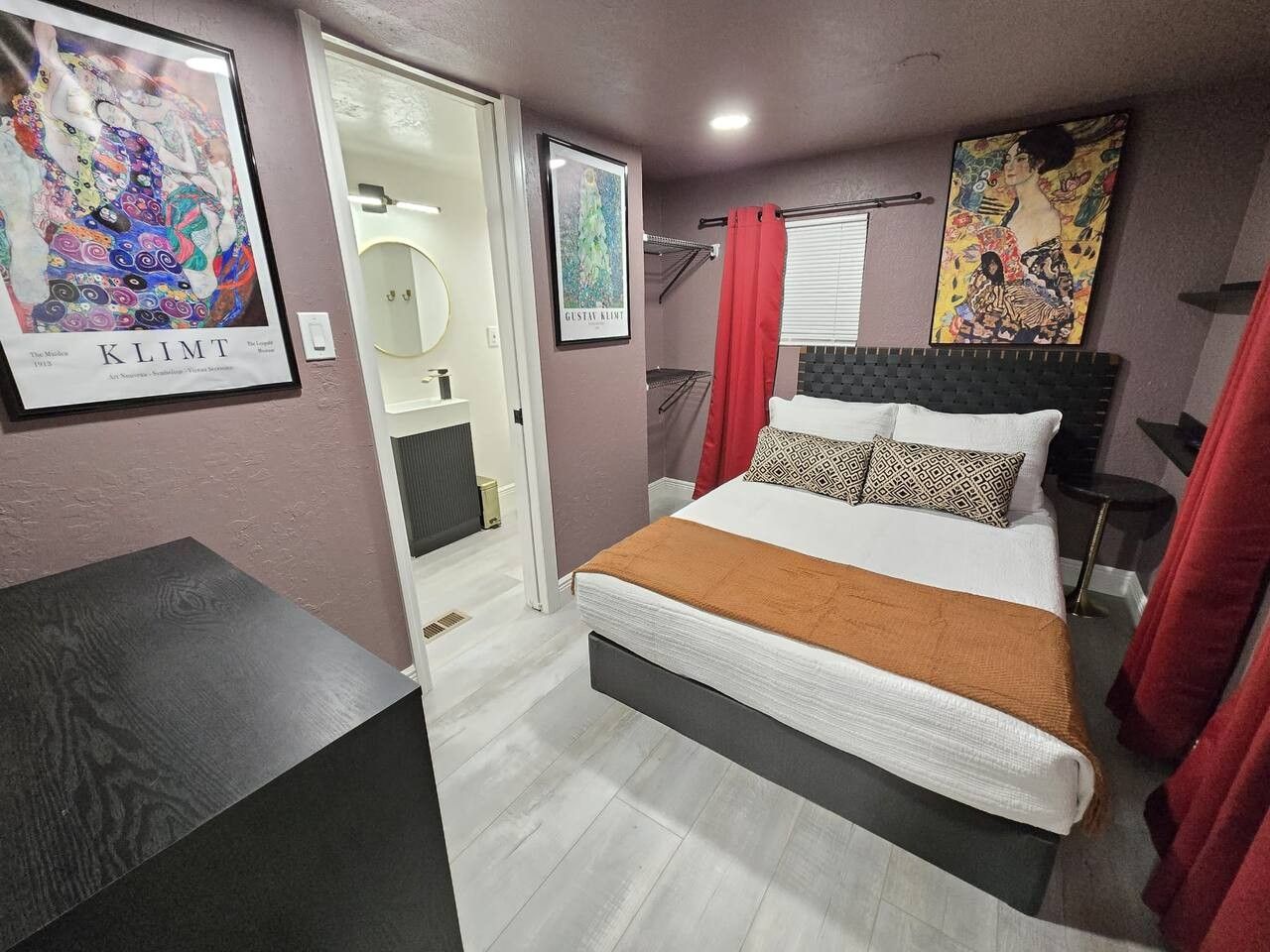 A bedroom with a bed, a bedside table, a red curtain, and artwork on the walls.