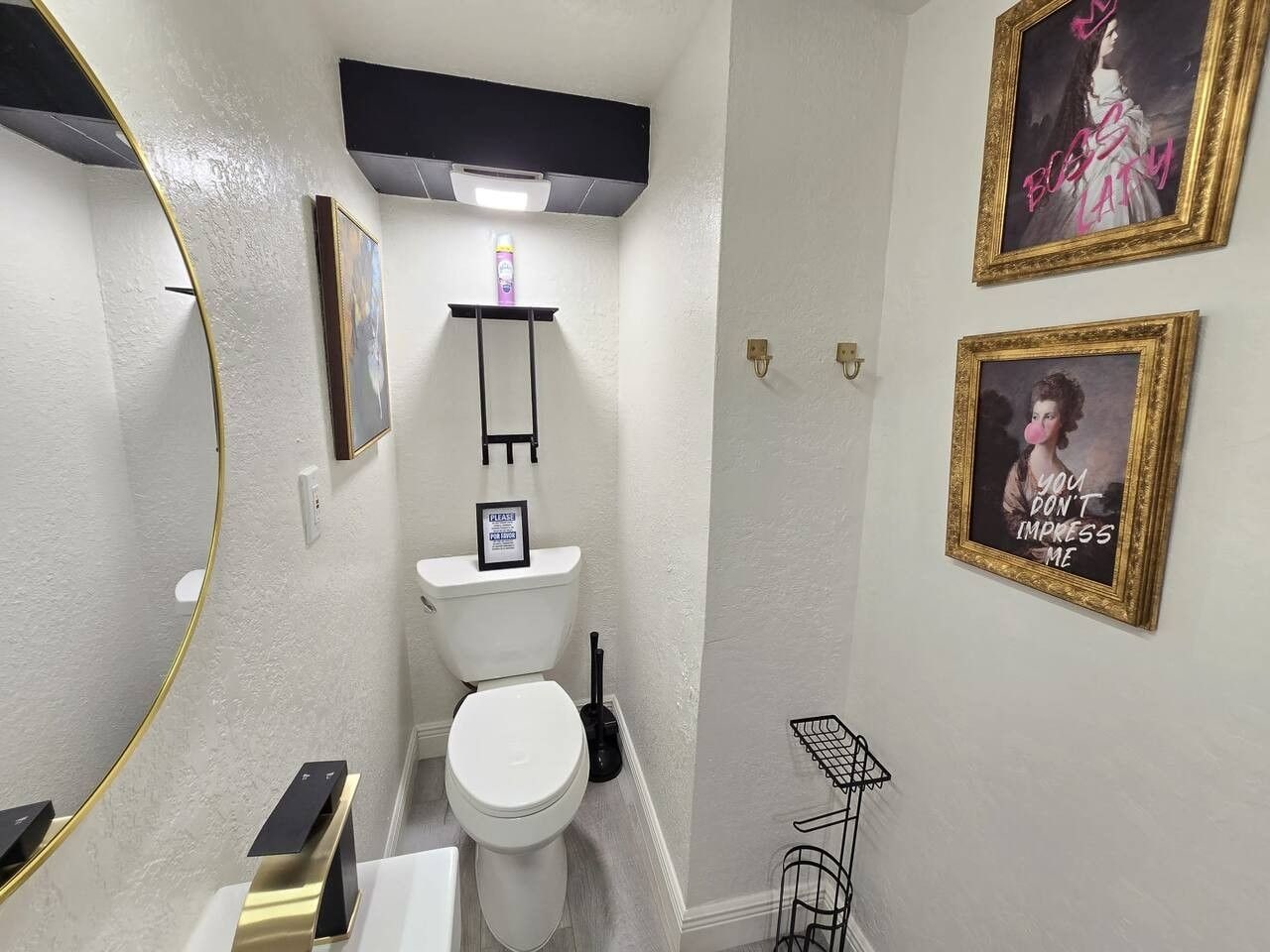 A modern bathroom with a toilet, a black shelf above the toilet, a framed picture on the wall, and a black floor lamp.