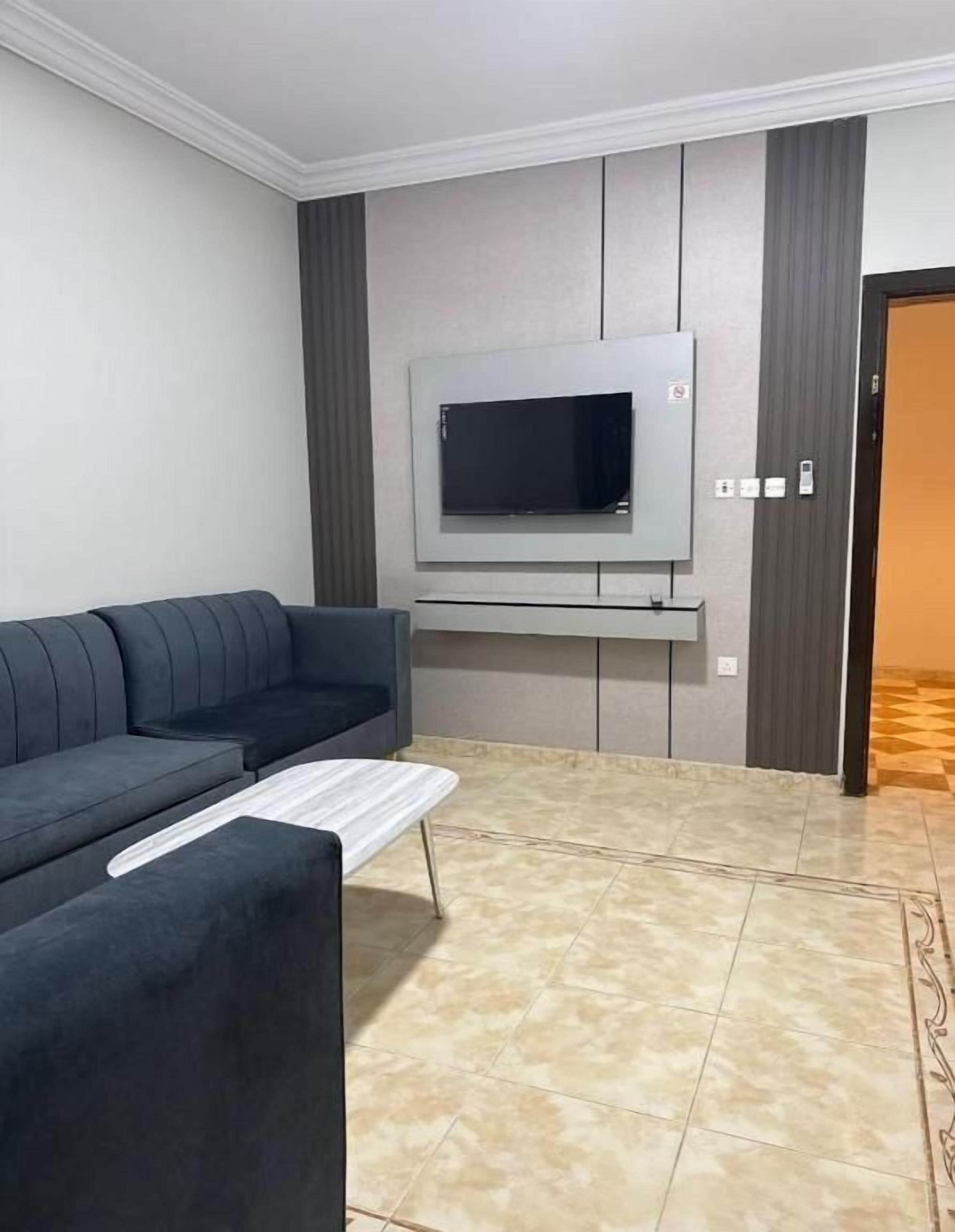 Una sala de estar con un sofá gris, un televisor de pantalla plana montado en la pared y un piso de baldosas.