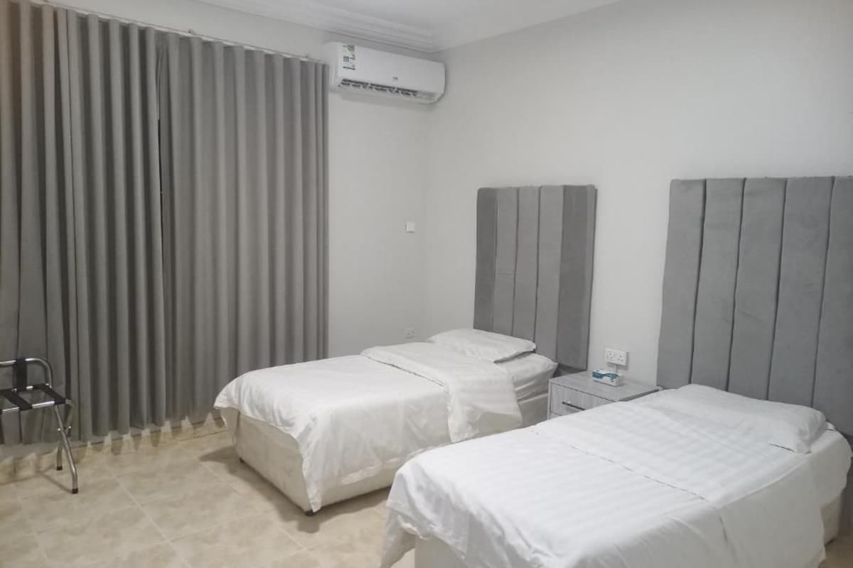 A hotel room with two beds, a chair, an air conditioning unit, and a nightstand.