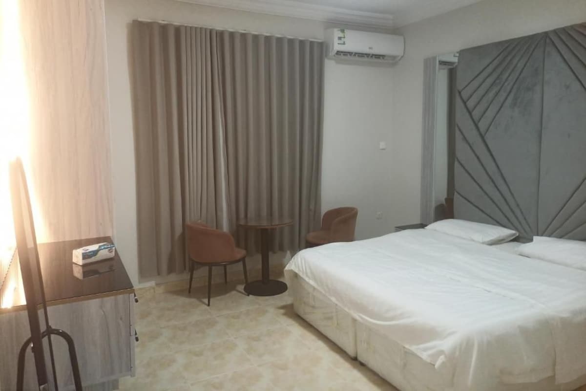 A hotel room with a bed, two chairs, a small table, an air conditioning unit, and a curtain.
