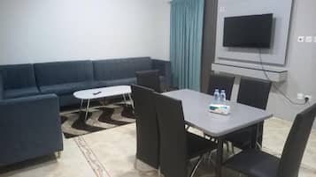 A modern living room with a sofa, a dining table with chairs, a flat-screen TV mounted on the wall, and a patterned rug.