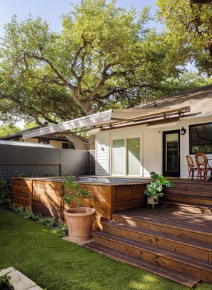 Een modern huis met een houten terras, een grote boom en een patio met stoelen.