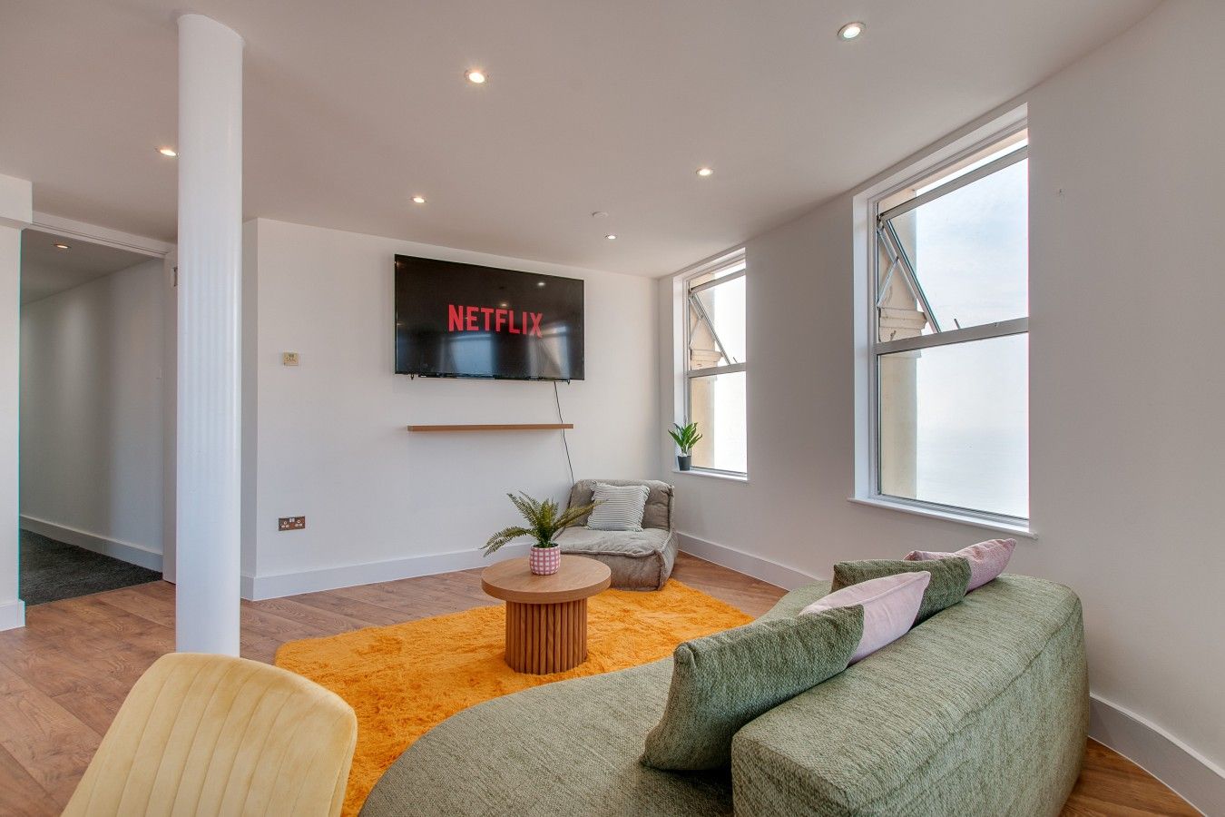 A modern living room with a flat-screen TV mounted on the wall, a round wooden coffee table, a green sofa with cushions, and a yellow chair.