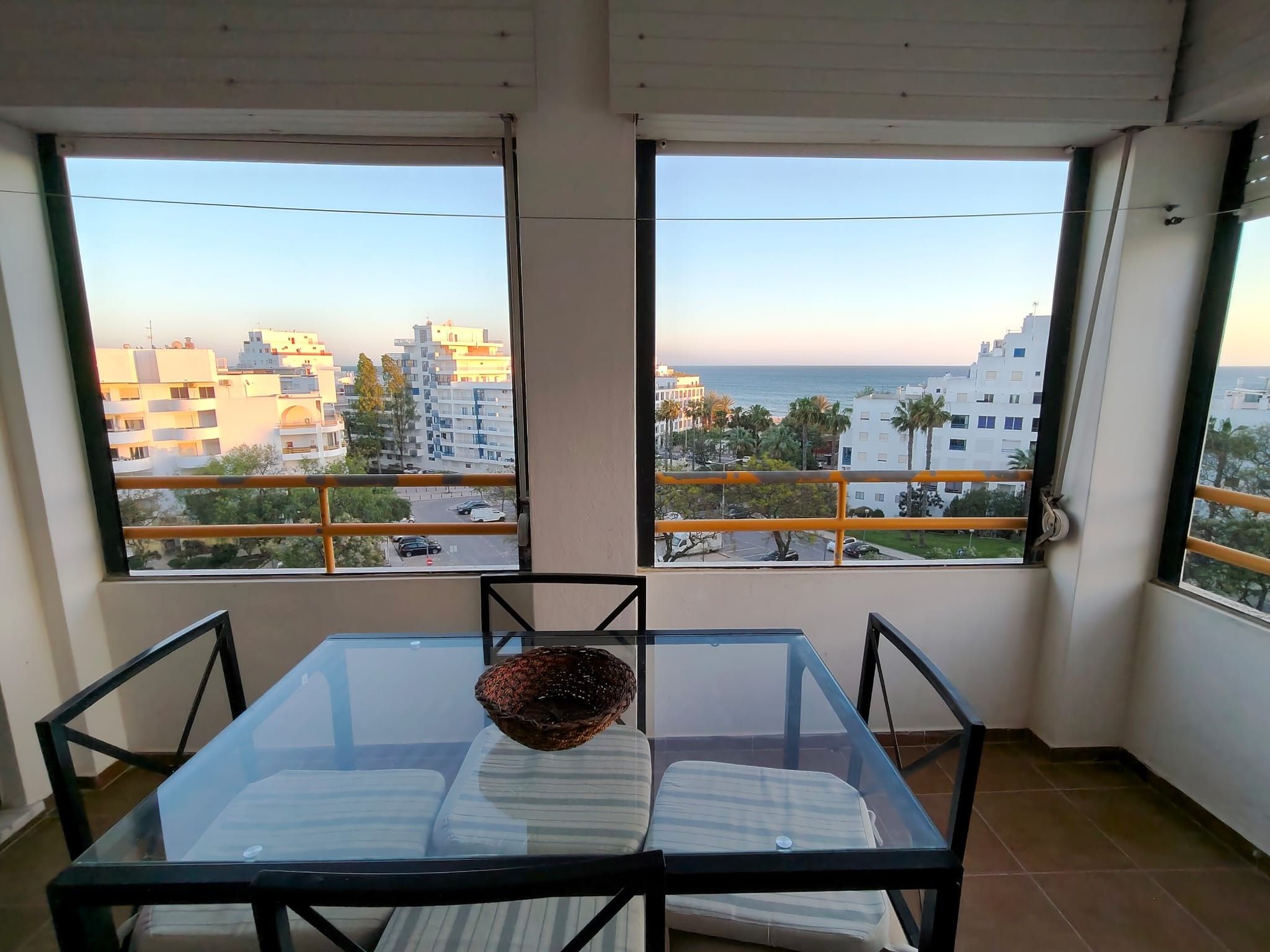 A balcony with a glass table and chairs, overlooking a cityscape with buildings and a clear sky.