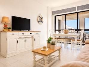 A modern living room with a flat-screen TV, white cabinetry, a small wooden table with a plant, and a view of the sea through large windows. - Apartment 'Mar Azul' with sea view, balcony and Wi-Fi (Quarteira)