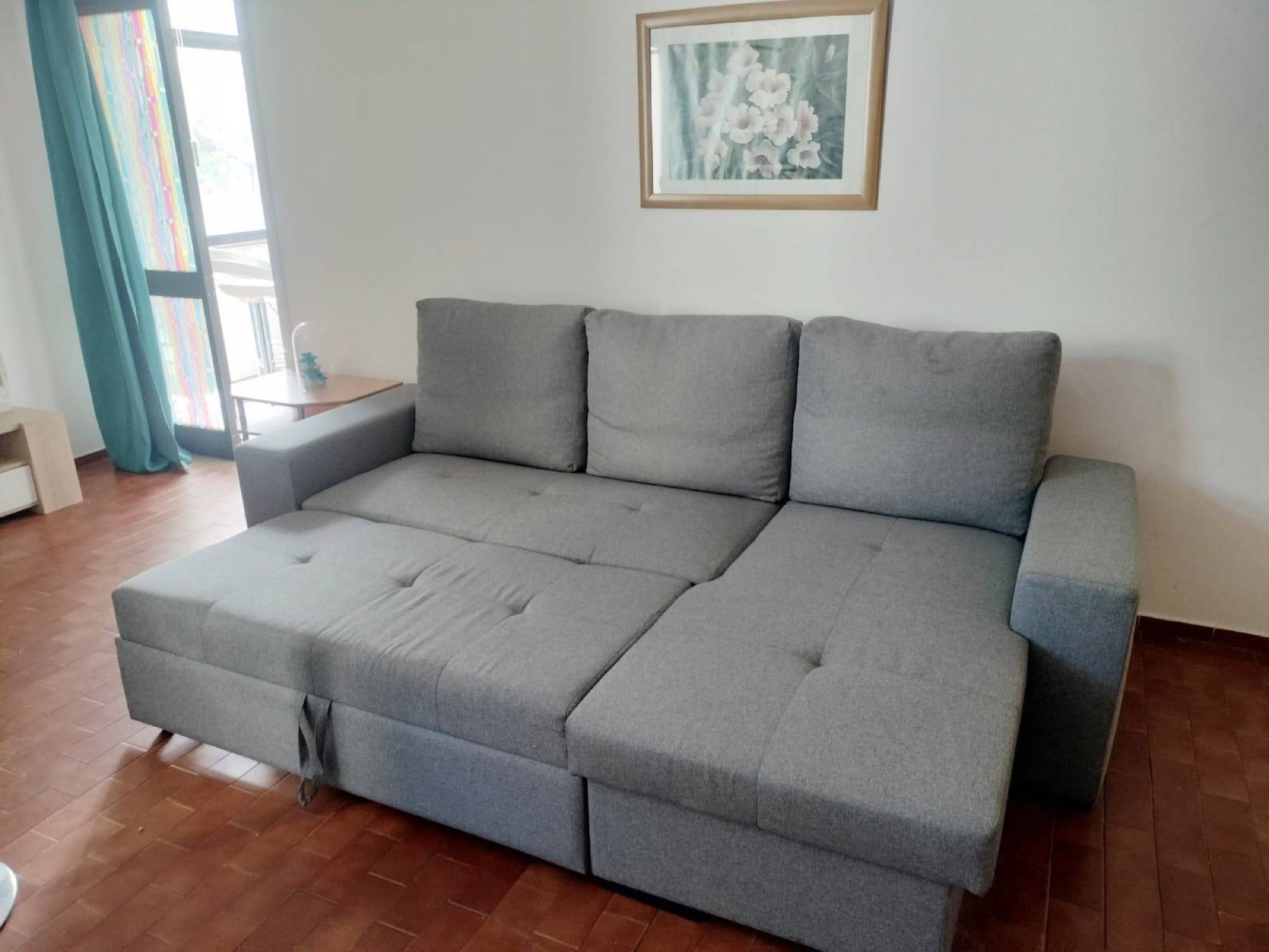 A grey sectional sofa with a wooden frame, a small side table, and a window with curtains.