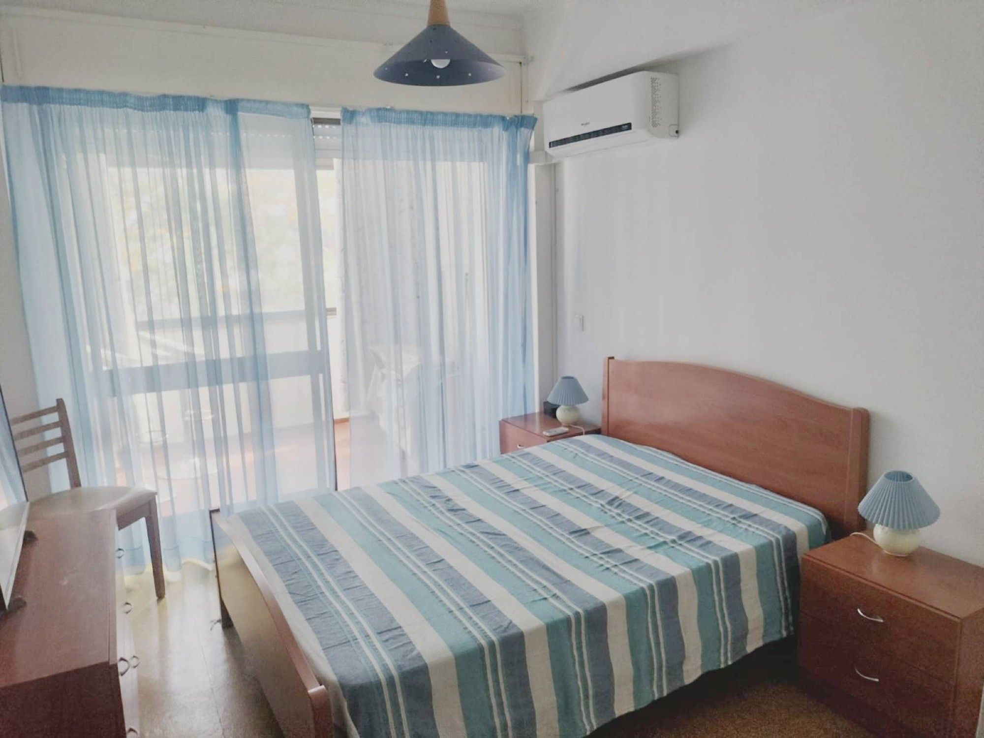A bedroom with a bed, a chair, a nightstand, a lamp, and a window with curtains.