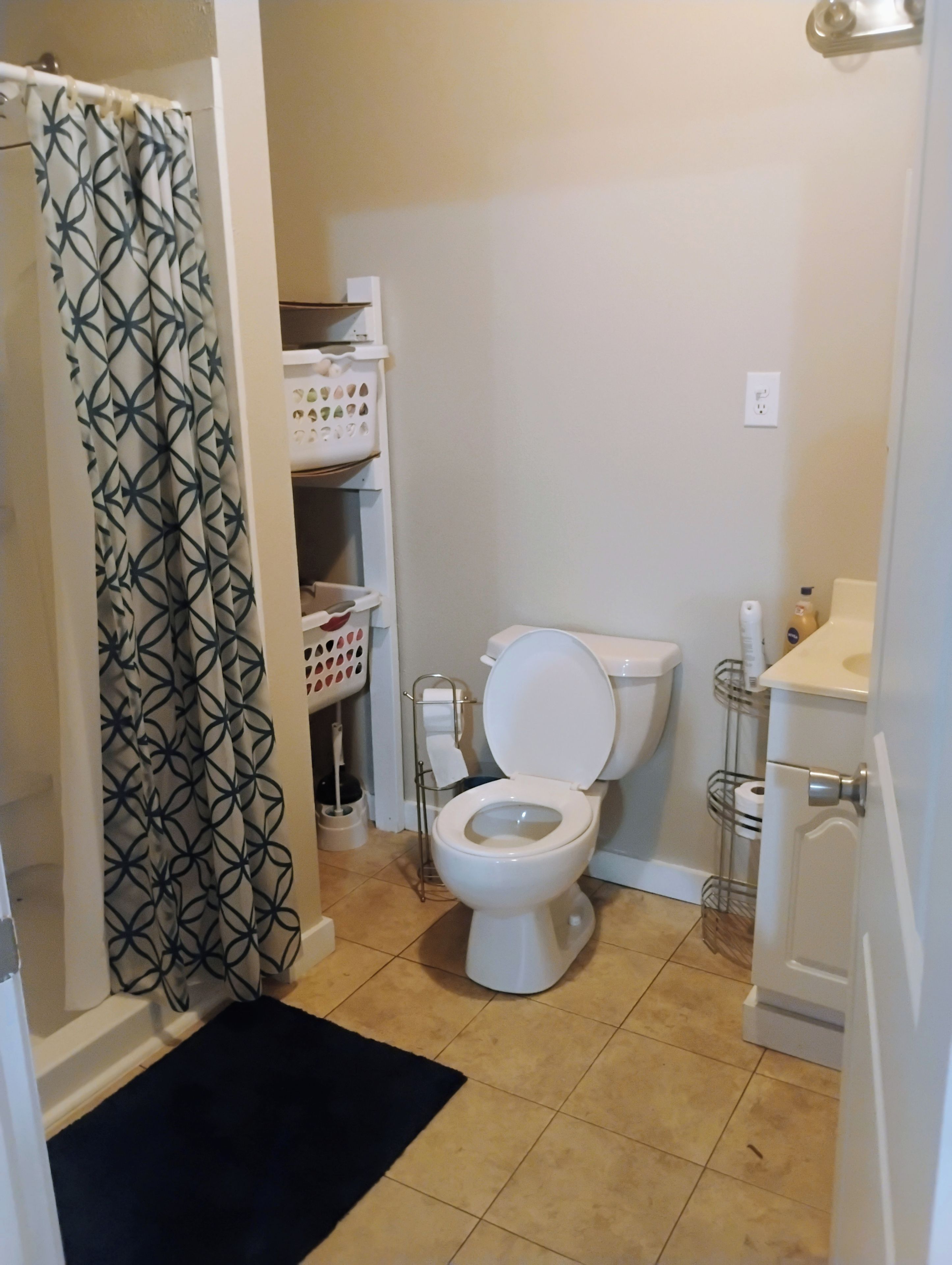 Une salle de bain avec des toilettes, un rideau de douche, une étagère contenant des articles de toilette et un tapis noir.
