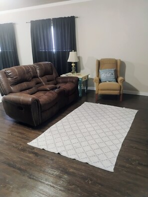 A living room with a brown leather sofa, a beige armchair, a patterned rug, a small table with a lamp, and a window with dark curtains. - Spacious studio apartment centrally located in Perry (Perry)