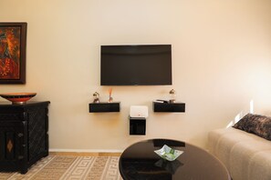 Una sala de estar con un televisor de pantalla plana montado en la pared, una mesa de centro negra, un sofá color beige y una alfombra estampada.