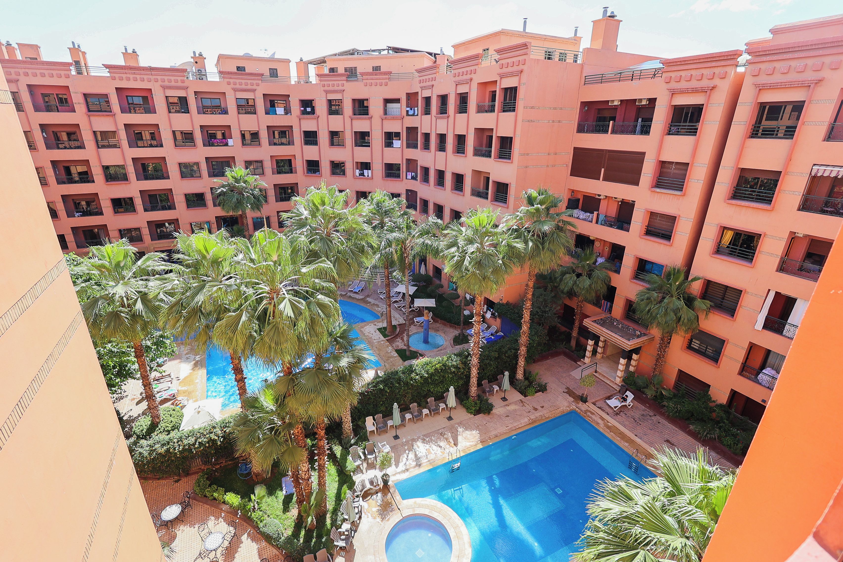 A multi-story hotel with a central courtyard featuring a swimming pool, palm trees, and lounge chairs.