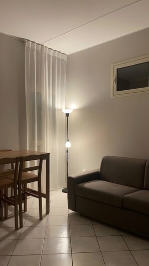 Ein Wohnzimmer mit einer Couch, einem Esstisch mit Stühlen, einer Stehlampe und einem Fenster mit Vorhängen.