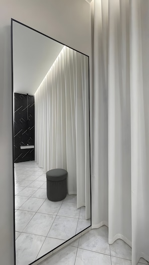 Ein modernes Badezimmer mit großem Spiegel, weißen Vorhängen, einem runden Hocker und Fliesenboden.