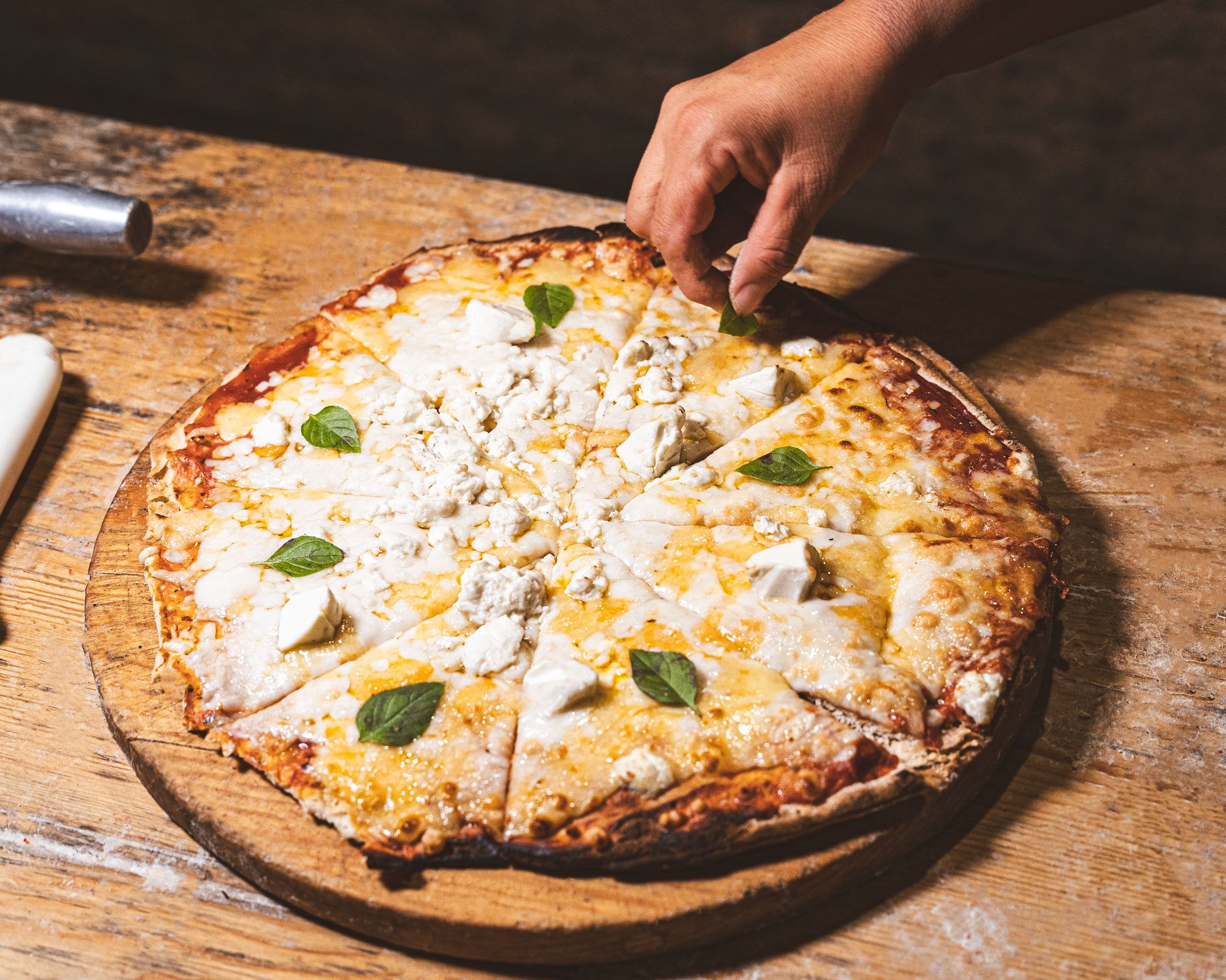 A pizza on a wooden board with a hand reaching for a slice.