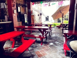 Un espacio de comedor al aire libre con mesas y bancos de madera roja, suelo de piedra y vista a un edificio al fondo.