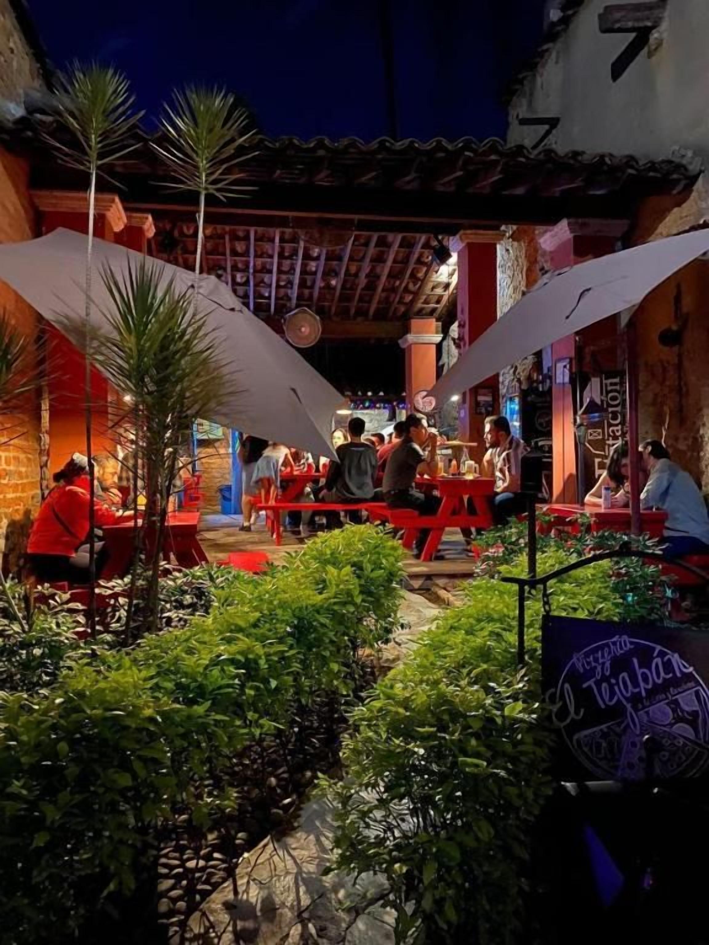Un área de comedor al aire libre con mesas y sillas, rodeada de follaje y un edificio con techo de tejas.