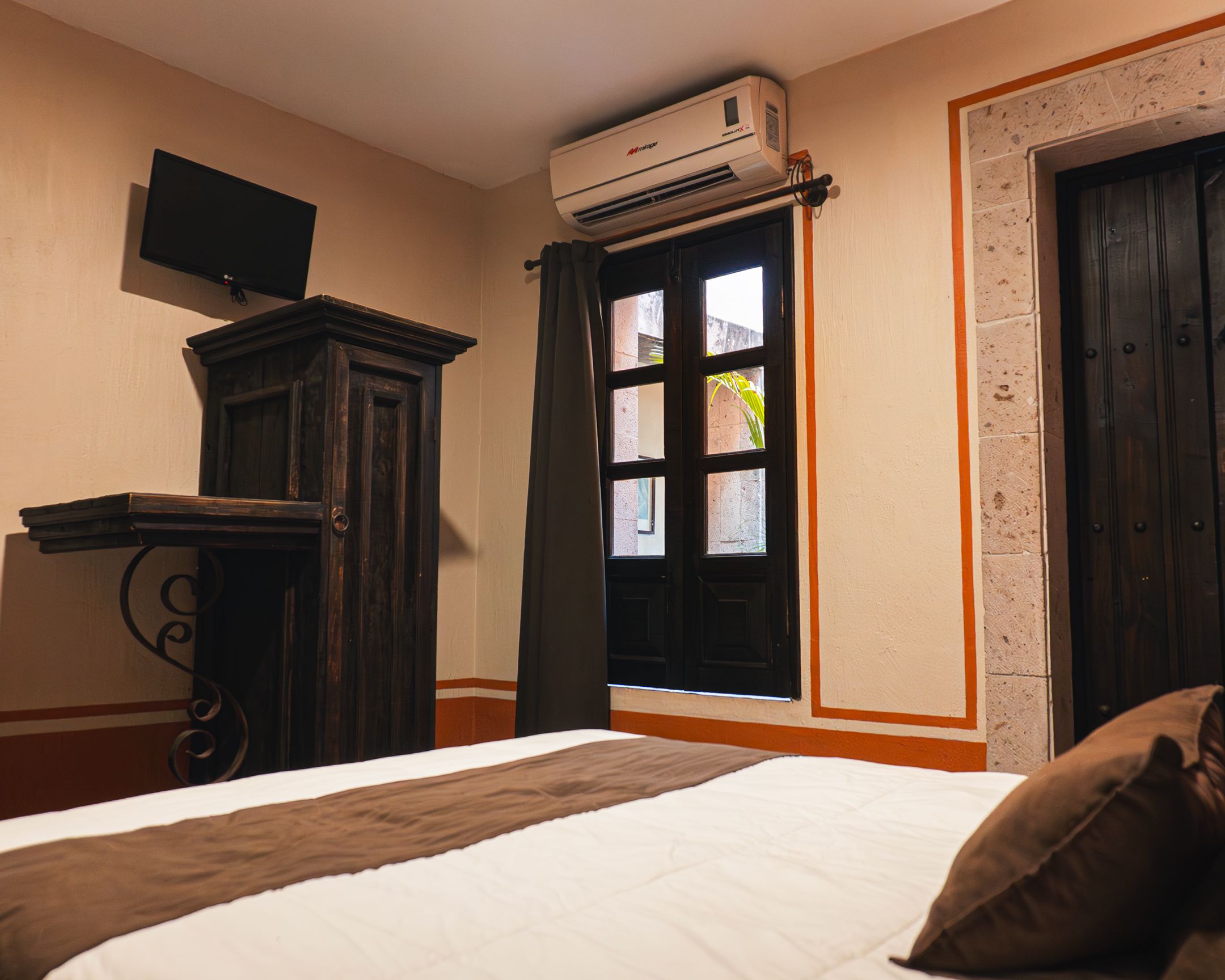A hotel room with a bed, a flat-screen TV mounted on a wooden cabinet, an air conditioning unit above a window, and a door with an orange trim.
