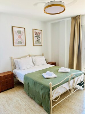 Un letto rifatto con una coperta verde e cuscini bianchi, un comodino in legno con un cassetto, una cornice con un'immagine appesa al muro e una finestra con tende color beige.