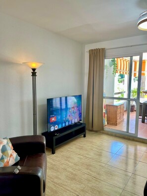 A living room with a flat-screen TV on a stand, a sofa, a floor lamp, and a sliding glass door leading to an outdoor area. - 2 bedroom Apartment sleeps 6, 5 min walk to the beach . (Benalmádena)